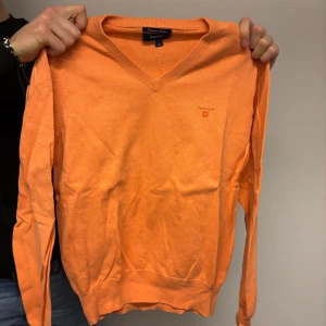Orange tröja från GANT - Säljer en snygg orange tröja från GANT. Tröjan har en v-ringad design och långa ärmar, perfekt för en stilren look. Den är i storlek M och har en diskret logga på bröstet.