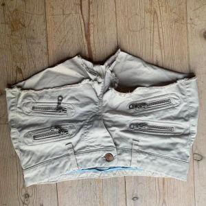 Beige lågmidjadeshorts med dragkedjor från Hunkydory - Snygga beige shorts från Hunkydory med flera dragkedjor som detaljer. De har en väldigt lågmidjad design och en knapp framtill. Perfekta för en cool och avslappnad stil.