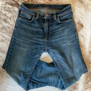 Blå jeans från Nudie Jeans - Snygga blå jeans från Nudie Jeans med klassisk femficksdesign och orange sömmar på bakfickorna. De har en knappgylf och är tillverkade i slitstarkt denim. Perfekta för en avslappnad stil.