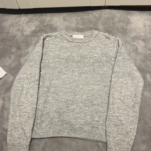 Grå stickad tröja med pärlor - Säljer en stilren grå stickad tröja dekorerad med pärlor på framsidan. Tröjan har långa ärmar och ribbstickade muddar vid ärmslut och nederkant. Perfekt för kyliga dagar!