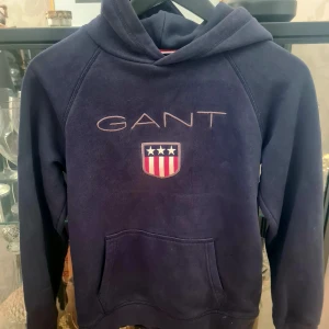 Mörkblå hoodie från GANT - Säljer en snygg mörkblå hoodie från GANT med deras ikoniska logga och sköld på bröstet. Tröjan har en klassisk känguruficka och en bekväm huva. Perfekt för en avslappnad stil.