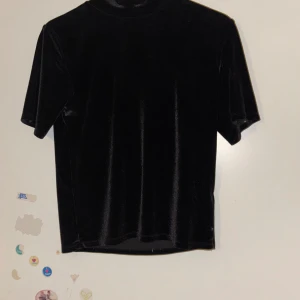 Svart sammetstopp från NA-KD - Säljer en elegant svart sammetstopp från NA-KD. Toppen har korta ärmar och en mjuk, glansig finish som ger en lyxig känsla. Perfekt för att höja vilken outfit som helst.