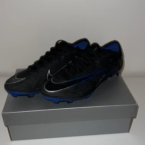 Nike air zoom mecurial vapor 15 elite FG - Snygga fotbollsskor från Nike i svart med blå och silver detaljer. Lite använda, fixat nya skosnören. Säljer dem på grund av att dom inte passar mig längre. Kom gärna med bud!