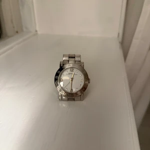 Silverfärgad klocka från Marc Jacobs - Säljer en fin Marc Jacobs damklocka. Klockan har en silverfärgad boett och armband. Urtavlan är vit med silverfärgade detaljer och strass stenar. Nypris 1800kr. Finns delar kvar för att ändra storlek.