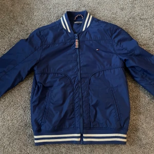 Blå bomberjacka från Tommy Hilfiger - Snygg blå bomberjacka från Tommy Hilfiger med dragkedja och ribbade muddar i vitt och blått. Jackan har en klassisk design med två fickor framtill och en liten logotyp på bröstet. Perfekt för en stilren look.