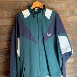 Grön och vit vindjacka från Reebok - Säljer en retro grön och vit vindjacka från Reebok med dragkedja och coola geometriska mönster. Jackan har långa ärmar och en hög krage. Perfekt för en sportig look eller en avslappnad dag ute.