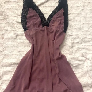 Lila topp/ klänning med spetsdetaljer - Säljer en elegant lila topp/  klänning med svarta spetsdetaljer vid axlarna. Klänningen har en v-ringad design och är perfekt för en stilfull look. Materialet är mjukt och följsamt.