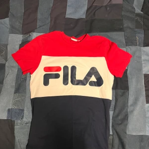 Flerfärgad t-shirt från FILA - Säljer en snygg flerfärgad t-shirt från FILA i rött, beige och svart. T-shirten har korta ärmar och ett stort FILA-tryck på framsidan. Perfekt för en sportig look eller avslappnad stil. Passar bra till jeans eller shorts!