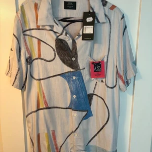 Mönstrad kortärmad skjorta från Neu Denim - Säljer en unik kortärmad skjorta från Neu Denim med abstrakt mönster i flera färger. Skjortan har en klassisk krage och knappar framtill. Perfekt för dig som vill sticka ut med en konstnärlig stil.