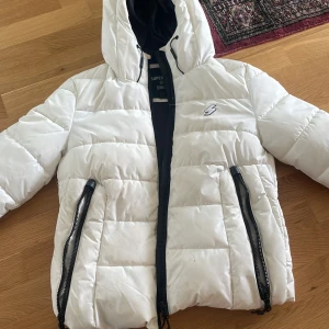 Vit pufferjacka från Superdry - Säljer en snygg vit pufferjacka från Superdry med svart dragkedja och huva. Jackan har en cool blixtdetalj på bröstet och är perfekt för kyligare dagar. Den har långa ärmar och praktiska fickor med dragkedja.