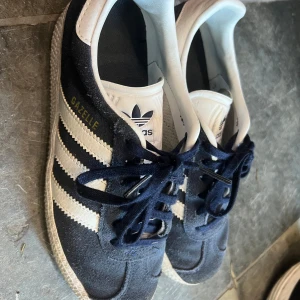 Adidas Gazelle sneakers - Snygga Adidas Gazelle sneakers i mörkblå mocka med vita detaljer och snörning. De har den klassiska tre-randiga designen på sidorna och en vit sula. Perfekta för en stilren och sportig look.