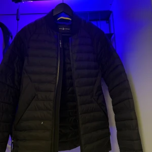 Svart dunjacka från sail racing - Säljer en stilren svart dunjacka från sail racing. Jackan har en dragkedja framtill och är quiltad för extra värme. Perfekt för kyligare dagar och har en modern look. Passar bra till både vardag och mer uppklädda tillfällen.