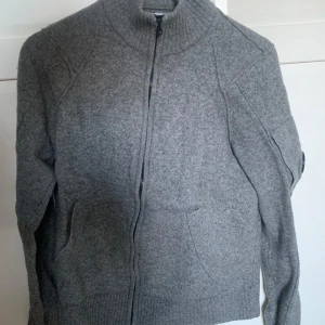 Grå stickad tröja från C.P. Company - Säljer en stilren grå stickad tröja från C.P. Company med dragkedja framtill. Tröjan har långa ärmar och en ribbad krage samt muddar. Perfekt för kyligare dagar.