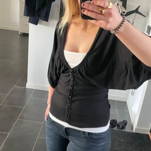 Svart kofta med knappar - Snygg svart kofta med knappar framtill och en djup v-ringning. Koftan har korta ärmar och en figurnära passform som framhäver midjan. 