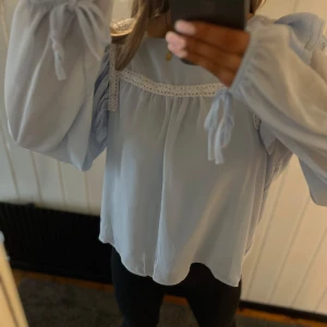 Ljusblå blus med volanger - En fin blus med en ljusblå färg samt vit, ger en booho stil. Den har volanger och knyten vid enden av armen vilket ger en söt stil på tröja🤩
