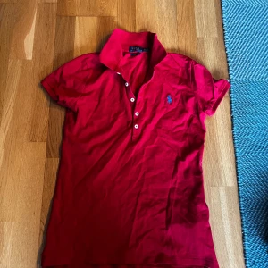 Röd pikétröja från Ralph Lauren - Säljer en klassisk röd pikétröja från Ralph Lauren med korta ärmar och knappar framtill. Tröjan har en broderad logga på bröstet och är i slim fit-modell. Perfekt för en stilren look!