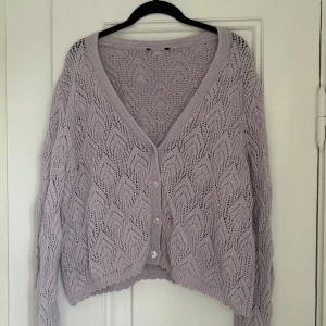 Ljuslila stickad kofta - Ljuslila stickad kofta med ett vackert hålmönster. Koftan har en v-ringad design och vackra knappar framtill. 