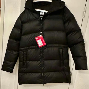 Svart dunjacka från Helly Hansen - Säljer en svart dunjacka från Helly Hansen med dragkedja och huva. Jackan har långa ärmar och är perfekt för kalla dagar. Den är designad för att hålla dig varm och bekväm.