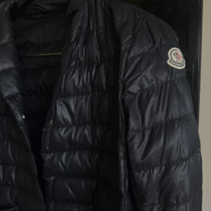 Svart dunjacka från Moncler - Snygg svart dunjacka från Moncler med quiltad design och lång ärm. Jackan har en klassisk krage och knäppning framtill. Perfekt för kyligare dagar. Kan inte garantera äktighet köpte second hand.
