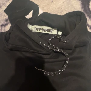Svart hoodie från Off-White - Säljer en svart hoodie från Off-White med ikoniska vita diagonala ränder på ärmarna och ryggen. Hoodien har en justerbar dragsko i huvan och en stor logotyp på baksidan. Perfekt för en stilren och modern look. Nypris: 2900