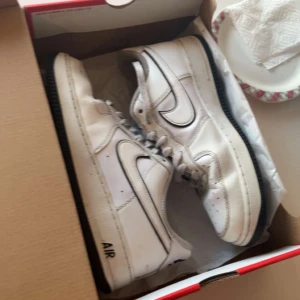 Vita Nike Air Force sneakers - Säljer ett par klassiska vita Nike Air Force sneakers med svart sula och detaljer. Skorna har den ikoniska swoosh-loggan i svart och texten 'AIR' på sidan. Perfekta för en stilren look.