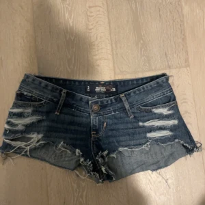 Blå jeansshorts med slitningar - Snygga blå jeansshorts från hollister med coola slitningar och fransiga kanter. Perfekta för en avslappnad stil. De har en knapp och dragkedja framtill samt klassiska bakfickor med dekorativa sömmar. Jag har sytt om de till lågmidjade de kan därför vara små i storleken då jag flyttat ner midjebandet. 