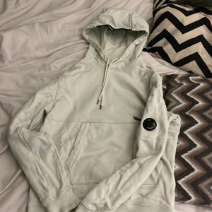 Ljusgrön hoodie från C.P. Company Storlek S - Säljer en stilren ljusgrön hoodie från C.P. Company. Den har en klassisk design med huva och dragsko samt en ficka framtill. En rund detalj på ärmen ger en unik touch. Perfekt för en avslappnad stil.