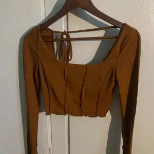 Säljer en snygg brun ribbad topp med långa ärmar och en croppad passform. Toppen har en unik design med knytning i ryggen och en fyrkantig halsringning. Perfekt för att ge din outfit en trendig touch!