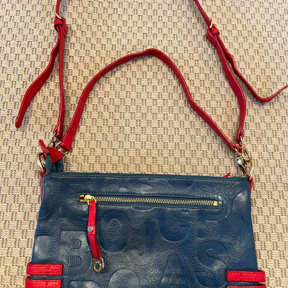 Cool blå väska med röda axelband och detaljer från Marc Jacobs. Mässingsspännen och dubbla axelband i olika längder. 1 mittenfack med magnetknapp. 2 stora fack med dragkedja och ett mindre fack med dragkedja på baksidan. Mått: 30x19 cm. Liten vit fläck på insidan av mittenfacket, se bild.. Laukut & Käsilaukut.