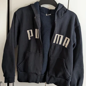 Svart hoodie från Puma - Säljer en svart hoodie från Puma med dragkedja och huva. Jackan har stora fickor framtill och ett mjukt foder. Perfekt för kyliga dagar. Sista biten av drag kedjan går trögt så jag säljer den billigt på grund av det. Den som köper kanske måste, troligen måste byta ut dragkedjan