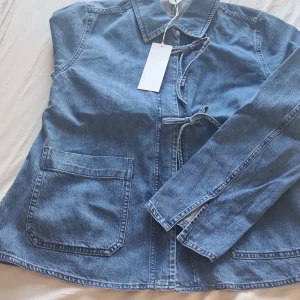 Blå jeansjacka med knytband Arket - Snygg blå jeansjacka med knytband i midjan och stora fickor framtill. Jackan har en klassisk krage och långa ärmar. Perfekt för en avslappnad stil.