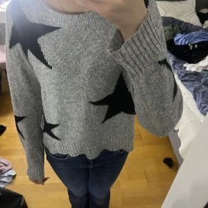 Grå stickad tröja med stjärnor - Säljer en grå stickad tröja med stora svarta stjärnor. Tröjan har en avslappnad passform och långa ärmar, perfekt för kyligare dagar. Den är både stilren och bekväm.