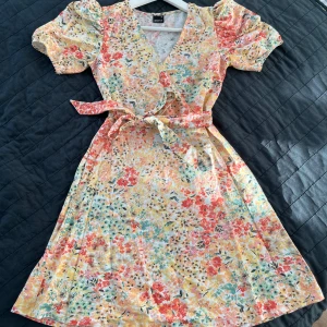 Blommig klänning med volangdetaljer - Charmig blommig klänning i ljusa toner med korta ärmar och v-ringning. Klänningen har volangdetaljer och ett knytband i midjan för en smickrande passform. Perfekt för sommarens alla tillfällen!