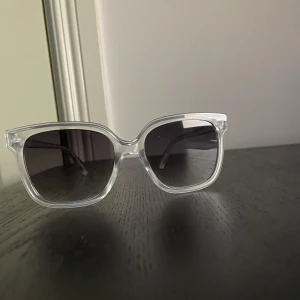 Transparenta John Henric solglasögon med svart toning - Snygga solglasögon med transparent båge och mörka linser. Perfekta för en stilren look. Passar både till stranden och stan.