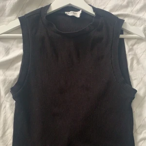 Svart ribbad ärmlös topp från Selma omari xNA-KD - Säljer en stilren svart ribbad topp från Selma omari xNA-KD. Den är ärmlös och har en rund halsringning, perfekt för lager-på-lager eller som den är. Passar till många olika stilar och tillfällen.