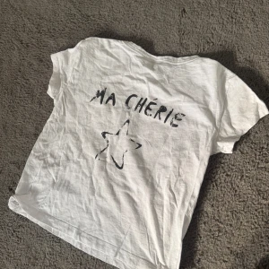 Vit t-shirt med tryck från Gina Tricot - Säljer en vit t-shirt från Gina Tricot med texten 'Ma Chérie' och en stjärna tryckt på framsidan. T-shirten har korta ärmar och är perfekt för en avslappnad stil. Passar bra till jeans eller shorts för en cool look.