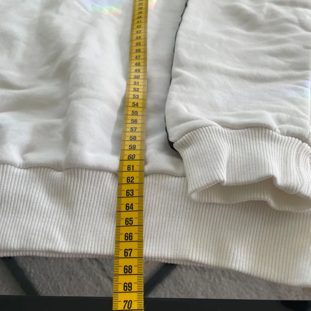 Säljer en stilren vit sweatshirt från Givenchy med svart text 'GIVENCHY PARIS' på framsidan. Tröjan har svarta detaljer längs ärmarna och ribbade muddar vid ärmslut och nederkant. Perfekt för en avslappnad och trendig look. Har tecken på använding och den är tillverkad i Turkiet.. Neuletakit & Villapaidat.