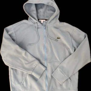 Ljusblå hoodie från Lacoste - Säljer en stilren ljusblå hoodie från Lacoste med dragkedja och klassisk passform. Tröjan har en broderad logotyp på bröstet och en praktisk huva med dragsko. Perfekt för en avslappnad look.