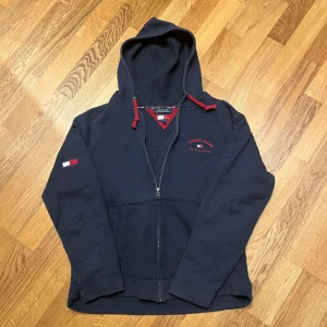 Mörkblå hoodie från Tommy Jeans (unknown slayer) - Säljer en snygg mörkblå hoodie från Tommy Jeans med dragkedja och huva. Den har röda detaljer och ett broderat märke på ärmen samt logotyp på bröstet. Perfekt för en avslappnad stil. Samma typ som unknown slayer och samma stil som Krobbe. 8/10 skick. Storlek: L men passar M.