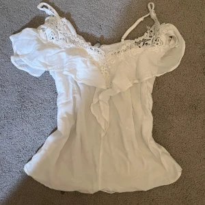 Vit spetsprydd linnetopp från Amisu - Säljer en söt vit linnetopp från Amisu med vackra spetsdetaljer och volang vid halsringningen. Toppen har justerbara axelband och är perfekt för varma sommardagar. Passar bra till både jeans och kjol.