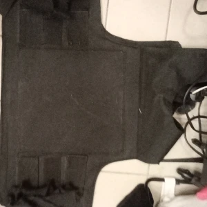 Svart kniv säker väst med kardborreband - Svart kinv säker väst med breda axelpartier och flera kardborreband framtill. Västen har en robust och funktionell design, perfekt för aktiviteter där extra stöd eller utrustning behövs. Materialet känns slitstarkt och västen har en enkel, stilren look.