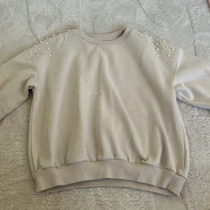 Beige tröja med pärldetaljer - Säljer en stilren beige tröja med pärldetaljer på axlarna. Tröjan har långa ärmar och en bekväm passform, perfekt för en avslappnad look. Den är gjord i ett mjukt material som känns skönt mot huden.