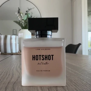 Hotshot but Hotter Eau de Parfum - Elegant Eau de Parfum från Linn Ahlborg, 'Hotshot but Hotter'. Flaskan har en stilren design med frostat glas och en svart fyrkantig kork. Perfekt för den som vill ha en doft som sticker ut.