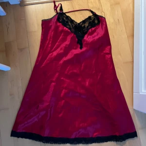 Röd satin slipklänning med spetsdetaljer - Säljer en röd satin slipklänning med svarta spetsdetaljer vid byst och nederkant. Klänningen har smala axelband och en v-ringad halsringning. Jättefin pjamas 