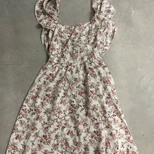 Blommig sommarklänning med volangdetaljer - Söt blommig klänning i vitt med rosa och gröna blommor. Klänningen har volangdetaljer vid axlarna och knytning i midjan. Perfekt för varma dagar och har en romantisk känsla.
