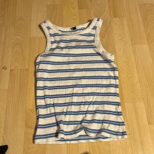 Vit och blårandig ribbad linnetopp - Säljer en vit ribbad linnetopp med blå horisontella ränder. Toppen har ärmlös design och rund halsringning. Perfekt för varma dagar eller som lager under andra plagg.
