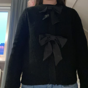 Svart kofta med rosetter - Säljer en svart kofta med två stora dekorativa rosetter framtill. Koftan har lång ärm och en mjuk, lite fluffig känsla. Perfekt för dig som vill ha en söt och stilren look.