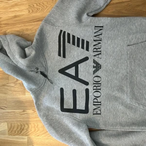  Emporio Armani - Säljer en grå hoodie från Emporio Armani med stor svart logga på framsidan. Tröjan har huva med dragsko och en stor magficka. Perfekt för en avslappnad och stilren look.