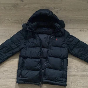 Svart dunjacka med huva - Säljer en svart dunjacka från Polo Ralph Lauren med huva och dragkedja. Jackan har långa ärmar och en broderad logotyp på bröstet. Perfekt för kalla dagar.