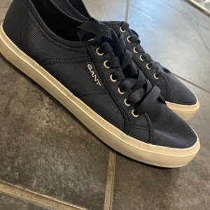Marinblå sneakers från Gant - Snygga marinblå sneakers från Gant med vita sulor och guldfärgade öljetter. Skorna har klassisk snörning och Gant-logga på sidan. Perfekta för en stilren och avslappnad look. De är nästintill oanvända